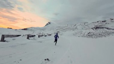 Arka planda karlı dağlarla birlikte koşan genç bir adamın hava aracı görüntüsü. Sağlıklı koşu ve egzersiz konsepti, yüksek hızlı insansız hava aracı FVP atışı, yavaş çekim.
