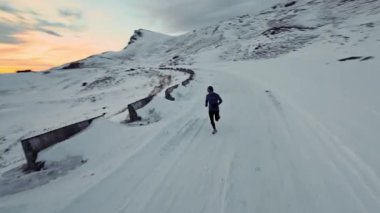 Arka planda karlı dağlarla birlikte koşan genç bir adamın hava aracı görüntüsü. Sağlıklı koşu ve egzersiz konsepti, yüksek hızlı insansız hava aracı FVP atışı, yavaş çekim.