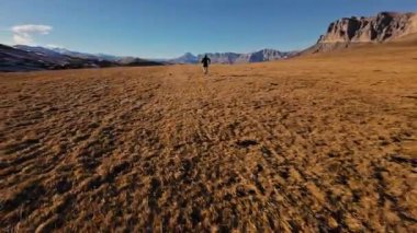 Sonbahar ya da kış aylarında dağ vadisinde arka planda dağlarla koşan spor giyimli FPV insansız hava aracı. Profesyonel koşucunun spor insansız hava aracı görünümü.