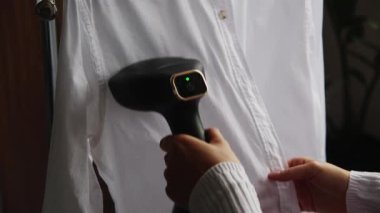 Yakın plan beyaz gömlekli, taşınabilir taşınabilir buharlı genç bir kadın. Ev hayatı ve rutin şeyler. Giysi bakımı.