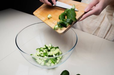 Salata hazırlamak için sebze doğramaya yakın çekim. Tahta kesme tahtasının üzerinde salatalık dilimleyip şeffaf bir kaseye döküyorum..