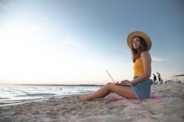Herhangi bir yerden çalış. Hasır şapkalı genç kadın güneş batarken tropik kumsalda otururken bilgisayarla çalışıyor..