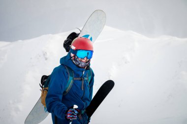 Miğferli snowboardda güzel bir gökyüzü ve güneşle dağın tepesinde duran kar sörfçüsü, güzel havalarda tatil köylerinde geçirdiği kış tatilleri için gülümsüyor ve mutlu..