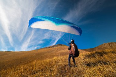 Bir paraglider yamaç paraşütü uçuşu için hazırlanır ya da çoktan tamamlamış ve çimenlerdeki bir tepeye konmuştur, canlı bir sonbahar manzarasıyla çevrili..