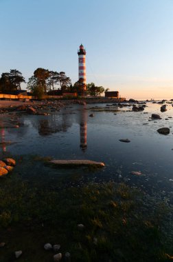 Sahildeki deniz fenerinde dramatik bir gün batımı. Ağaçların arasında beyaz ve kırmızı çizgili deniz feneri.
