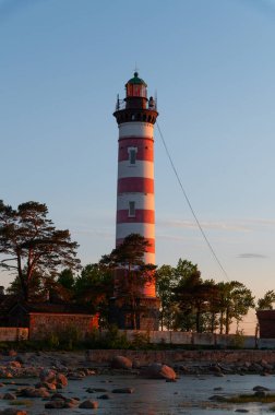 Sahildeki deniz fenerinde dramatik bir gün batımı. Ağaçların arasında beyaz ve kırmızı çizgili deniz feneri.