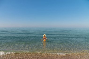 Sabahın erken saatlerinde sahilde bikinili ve şapkalı genç bir kadın, sakin denize doğru yürüyor..