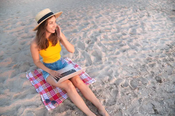 Kumsalda battaniye üzerinde otururken dizüstü bilgisayarda yazan ve denize bakan mutlu kadın. Uzak çalışma ya da öğrenim.