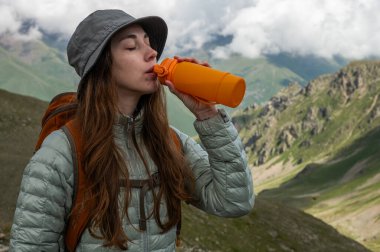 Ormanda yürürken sırt çantasıyla su içen çekici, mutlu, genç bir bayan yürüyüşçü. Yazın dağlarda tek başına zirveye çıktıktan sonra soğuk içecekle rehidrasyon..
