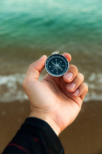 Pov of man hand holding navigational travel compass to find direction and next destination place. Concept of travel people and exploring. Beach and ocean in background.