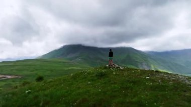 Bir kayanın tepesindeki bir adam ve yazın renkli bir gökyüzünde bulutun içindeki güzel bir dağ vadisi. Guy, sisli dağ sıraları, yeşil çimenler, güneş batarken beyaz gökyüzü. Dağlarda koşuyor..