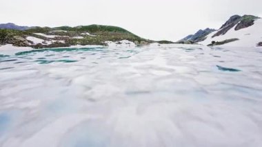 Fpv spor drone 'lu kadının, turkuaz yüksek dağ gölündeki kürek tahtasının üzerinde yüzen havadan görüntüsü. Yüksek kayalık tepeler ve karlı kıyılar.