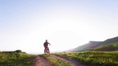 Miğferli ve maskeli genç bir sporcu gün batımında arka tekerlere binip pedal çeviriyor. Fpv insansız hava aracında hava görüntüsü.