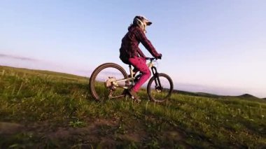 Hava görüntüsü yavaş çekim fpv kuadkopter drone görüntüsü günbatımında tepede bisiklet süren bir bisikletçiyi takiben. Arka planda destansı kayalık yeşil çimenler var. Kasklı ve maskeli profesyonel bisikletçi gün batımında dışarı çıkıyor.