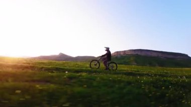 Miğferli ve maskeli genç bir sporcu gün batımında arka tekerlere binip pedal çeviriyor. Fpv insansız hava aracında hava görüntüsü