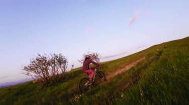 Hava görüntüsü fpv kuadkopter drone görüntüsü arka planda destansı kayalık yeşil çimlerle gün batımında tepede bisiklet süren bir bisikletçiyi takip ediyor. Kasklı ve maskeli profesyonel bisikletçi gün batımında dışarı çıkıyor.