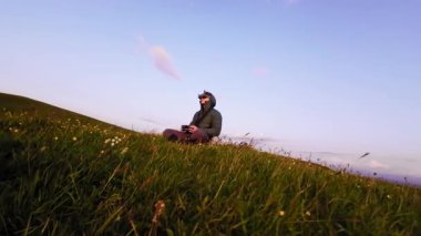 FPV gözlük takan beyaz bir adam gün batımında yeşil çimlerin üzerinde dağlık arazide oturuyor. Ağır çekim. Hava görünümü.