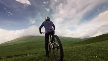 Hava fpv görüntüsü. Bisikletli kasklı genç adam dağın tepesinde dikiliyor. Bulutlu ve kayalık dağlı destansı bir arka plan. Kamera onun etrafında dönüyor. 4 bin. Yakın plan.