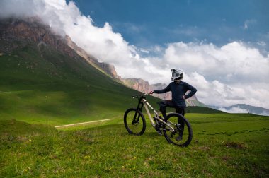Tatmin olmuş bir adam, kaskı ve gözlüğü içinde, dağları kaplayan bulutlarla dolu bir dağ bisikletiyle gökyüzünün altında duruyor. Sıra dışı spor.