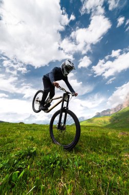 Bir dağ bisikletçisi, arka tekerleği ön tekerleğine dayanmış, yazın tarlalarda bir dağ manzarasına karşı manuel ya da tekerlek numarası yaparak havalanıyor..