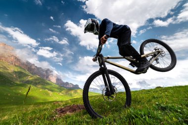 Tam yüzlü kask takan profesyonel bir adam yazın dağlarda bir tarlada ön teker numarası yapar. Tanınmayan bir sporcu elle bir numara gösterir..