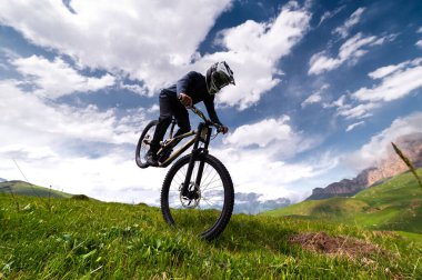 Tam yüzlü kask takan profesyonel bir adam yazın dağlarda bir tarlada ön teker numarası yapar. Tanınmayan bir sporcu elle bir numara gösterir..