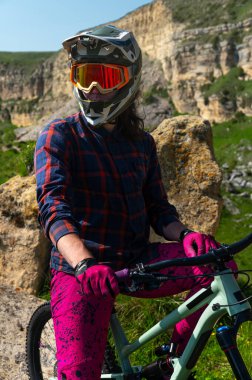 Koruyucu kasklı ve eldivenli, parlak elbiseli bir gencin portresi, yokuş aşağı bir bisiklette oturuyor ve uzaklığa bakıyor. Dağlarda aşırı dinlenme.