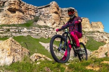 Koruyucu kasklı ve eldivenli, parlak elbiseli bir gencin portresi, yokuş aşağı bir bisiklette oturuyor ve uzaklığa bakıyor. Dağlarda aşırı dinlenme.