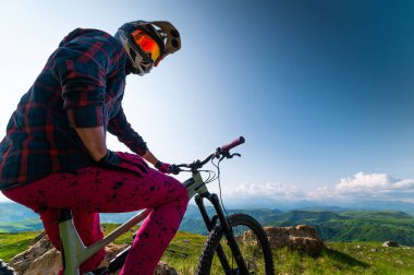 Pembe takım elbiseli bir bisikletçinin geniş açılı görüntüsü. Bisikletinin üstünde oturuyor ve kameraya bakıyor yemyeşil tepelerin ve kümülüs bulutlarının olduğu bir arka planda mavi gökyüzüne bakıyor..