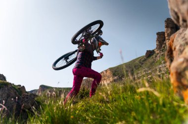 Bisiklet kıyafetli bir adam omuzunda dağ bisikleti taşıyarak bir patika boyunca yürür. Genç bir adam bisikletini alır ve dağlara taşır..