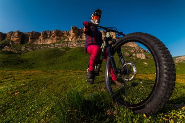 Öndeki tepeden aşağıya doğru inen bisiklet tekerleğinin geniş açılı görüntüsü, sporcunun güvenli bir şekilde bisiklette oturması. Arka planda keskin kayalık dağlar görülebilir..
