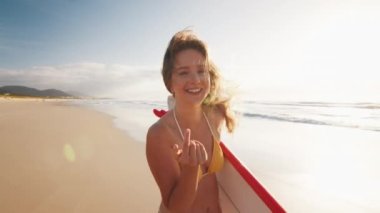 Seksi sörfçü. Bikinili genç mutlu kadın kumlu sahilde sörf tahtasıyla yürüyor.