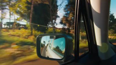 Hareket halindeki arabadan yol manzarası. Arabanın plakası asfalt yolda ilerliyor. Güneş ağaçların arasından parlıyor, hareket halindeki arabanın manzarası