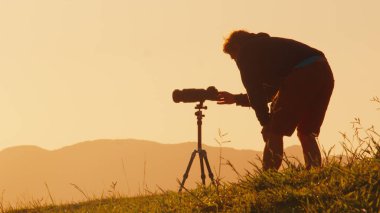 Tepedeki fotoğrafçı. Genç erkek fotoğrafçı, güneş doğarken dağların yeşil tepesindeki tripoda kamera ve telefoto lenslerle fotoğraf çekiyor.