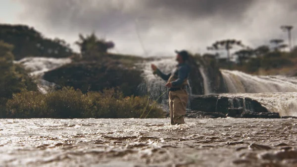 フライフィッシング 水夫の漁師は 背景に滝がある急速な濁った川でラインと釣りをキャストします 前景の水に焦点を当てる — ストック写真