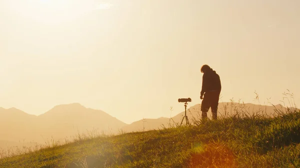 丘の上の写真家 若い男性写真家は日の出に山の緑の丘の上に三脚にセットされたカメラと望遠レンズで写真を撮ります — ストック写真