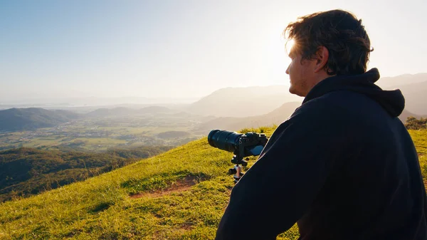 丘の上の写真家 若い男性写真家は日の出に山の緑の丘の上に三脚にセットされたカメラと望遠レンズで写真を撮ります — ストック写真
