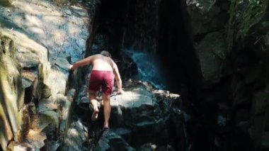 Genç adam duş almak ve şelalede dinlenmek için kayalık duvara tırmanıyor.