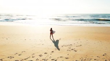 Seksi sörfçü havası. Bikinili genç sörfçü kadının kumlu mükemmel plajda sörf tahtasıyla yürüyüşünün havadan görüntüsü.