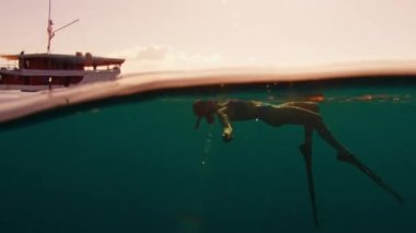 Serbest dalış yapan kadın gün batımında denizde yüzer ve arka planda tekne ile yüzeyin altında rahatlar ve yüzer.