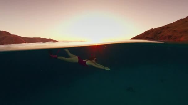 ピンクのスーツを着た女性が水中で泳ぎ 日没時にインドネシアのコモド国立公園の水中世界を探検する — ストック動画