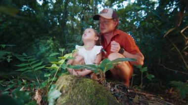 Aile bilimi. Çocuklu yetişkin bir adam tropikal ormanda oturur ve yerde koşan karıncaları seyreder. Baba kızına doğanın harikalarını gösterir.