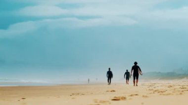 Sörf tahtalı sörfçüler arka planda sisli dağlarla sahilde yürürler.