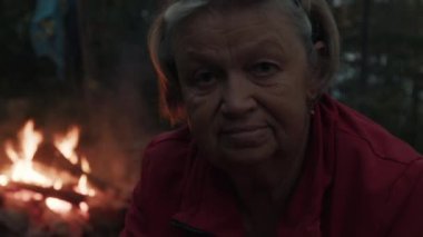 Arka planda yanan ateşle ormanda kamp yapan yaşlı kadının portresi.
