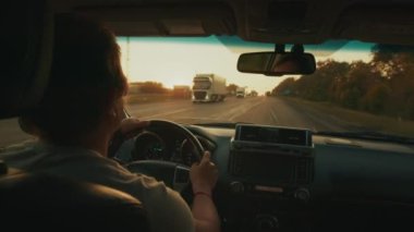 Gün batımında kamyonlarla dolu otoyolda ağır trafikte direksiyon sallayan kişi.