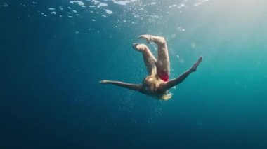 Kırmızı elbiseli genç kız kristal berrak tropikal denizde güvenli bir şekilde suya dalıyor ve yüzüyor. Suda güneş parlıyor.