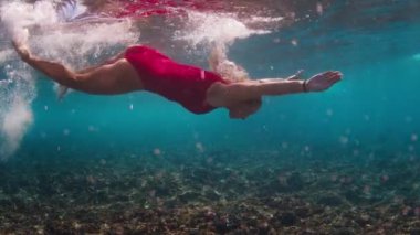 Kırmızı elbiseli genç kadın dalganın altına dalıyor ve sert deniz ortamında güvenli bir şekilde suyun altında yüzüyor.