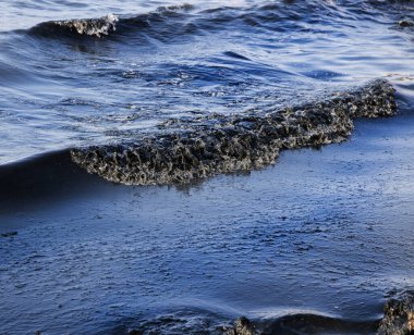 Kaya kıyısı, petrol sızıntısı gökkuşağı parıltısıyla kaplı karanlık dalgalarla vuruyor, çevresel felaketin sembolü ve petrol kirliliği sonrasında deniz kirliliğinin yol açtığı deniz yaşamına tehdit oluşturuyor.