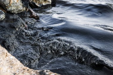 Kaya kıyısı, petrol sızıntısı gökkuşağı parıltısıyla kaplı karanlık dalgalarla vuruyor, çevresel felaketin sembolü ve petrol kirliliği sonrasında deniz kirliliğinin yol açtığı deniz yaşamına tehdit oluşturuyor.