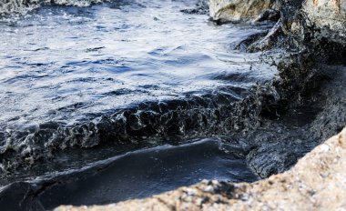 Kaya kıyısı, petrol sızıntısı gökkuşağı parıltısıyla kaplı karanlık dalgalarla vuruyor, çevresel felaketin sembolü ve petrol kirliliği sonrasında deniz kirliliğinin yol açtığı deniz yaşamına tehdit oluşturuyor.